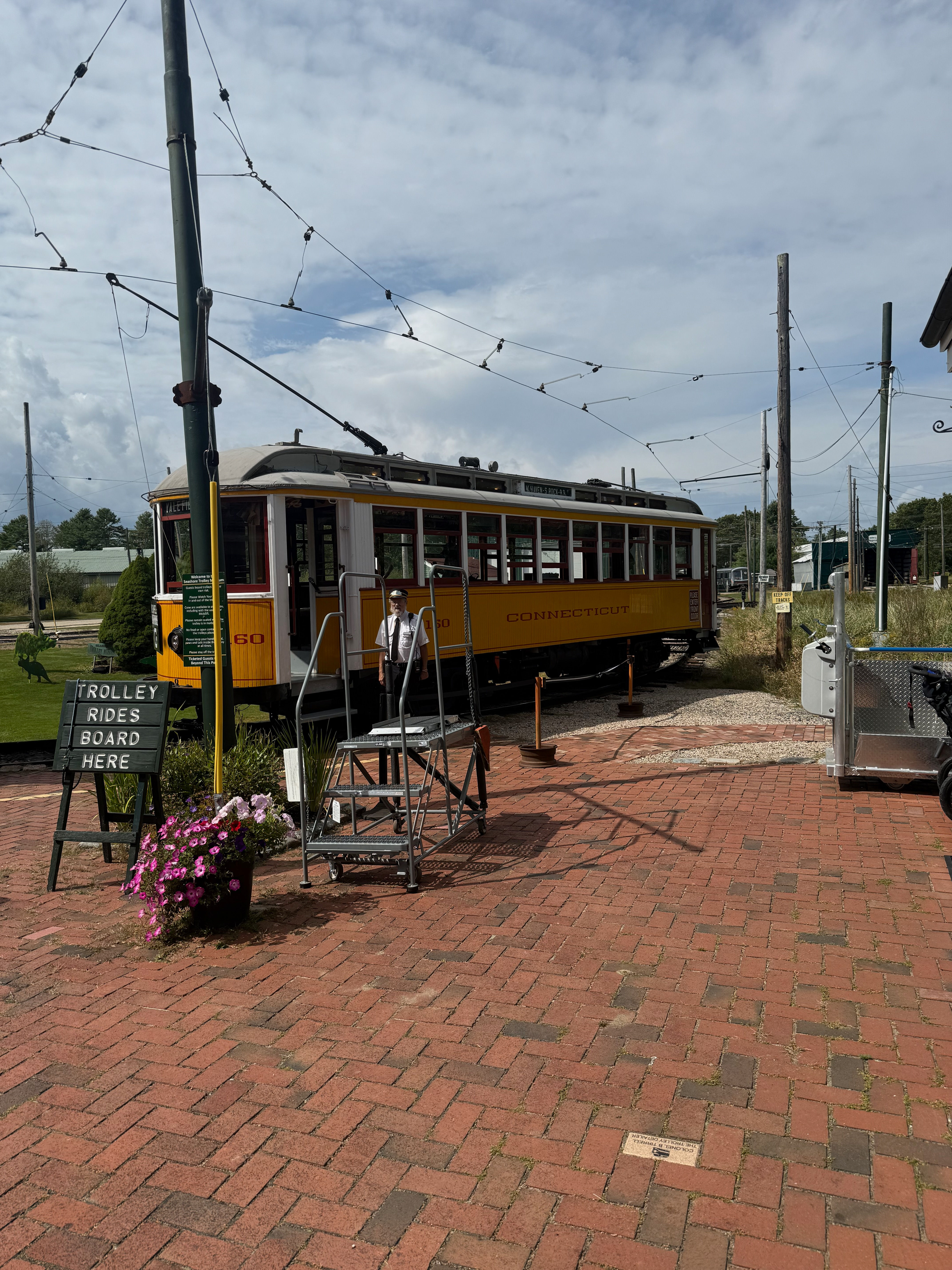 A vintage yellow and white trolley car from “THE CONNECTICUT CO.” is stopped on the tracks next to a brick platform. A small A-frame sign reads “TROLLEY RIDES BOARD HERE” and a metal step stool provides access to the trolley’s open door.