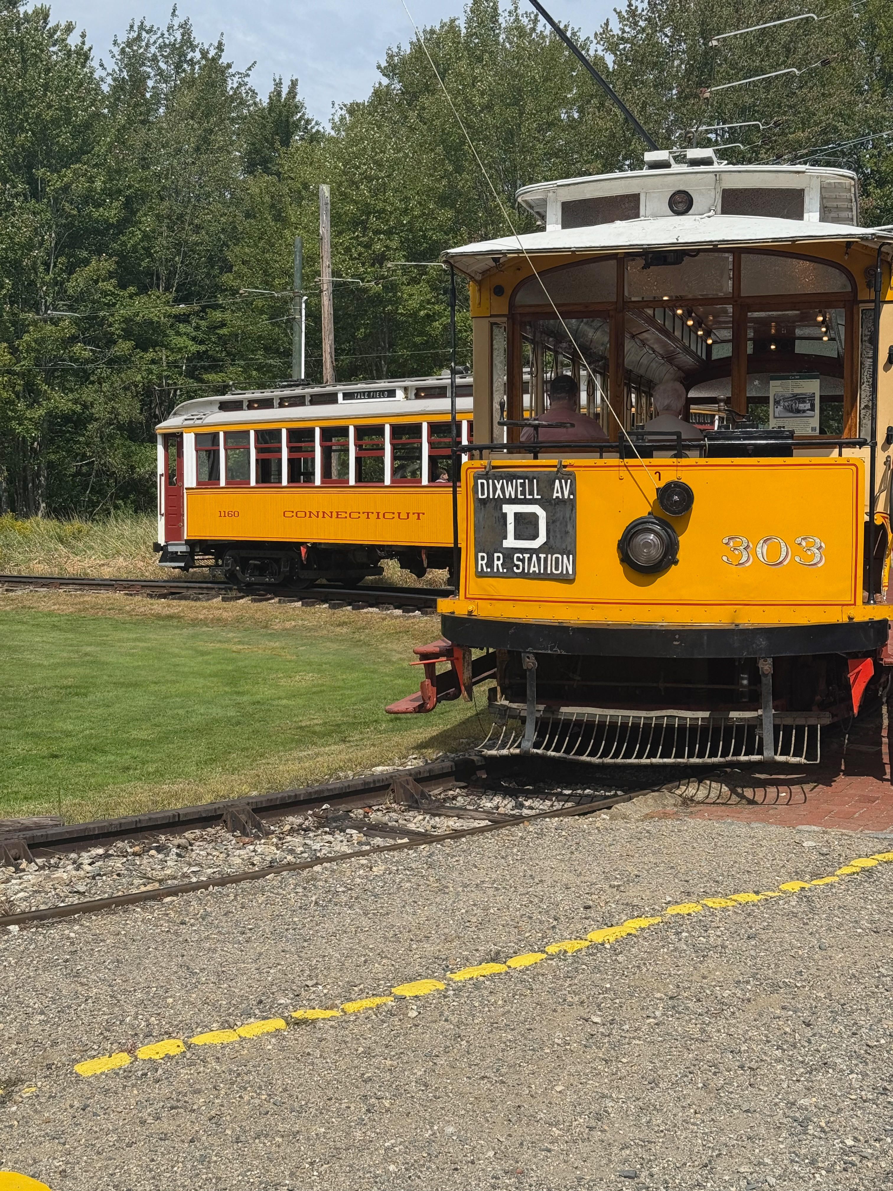 A close-up of the front of a vintage yellow trolley, number 303. The destination sign reads “DIXWELL AV. R.R. STATION”. In the background on a parallel track, another vintage trolley is visible among green trees.