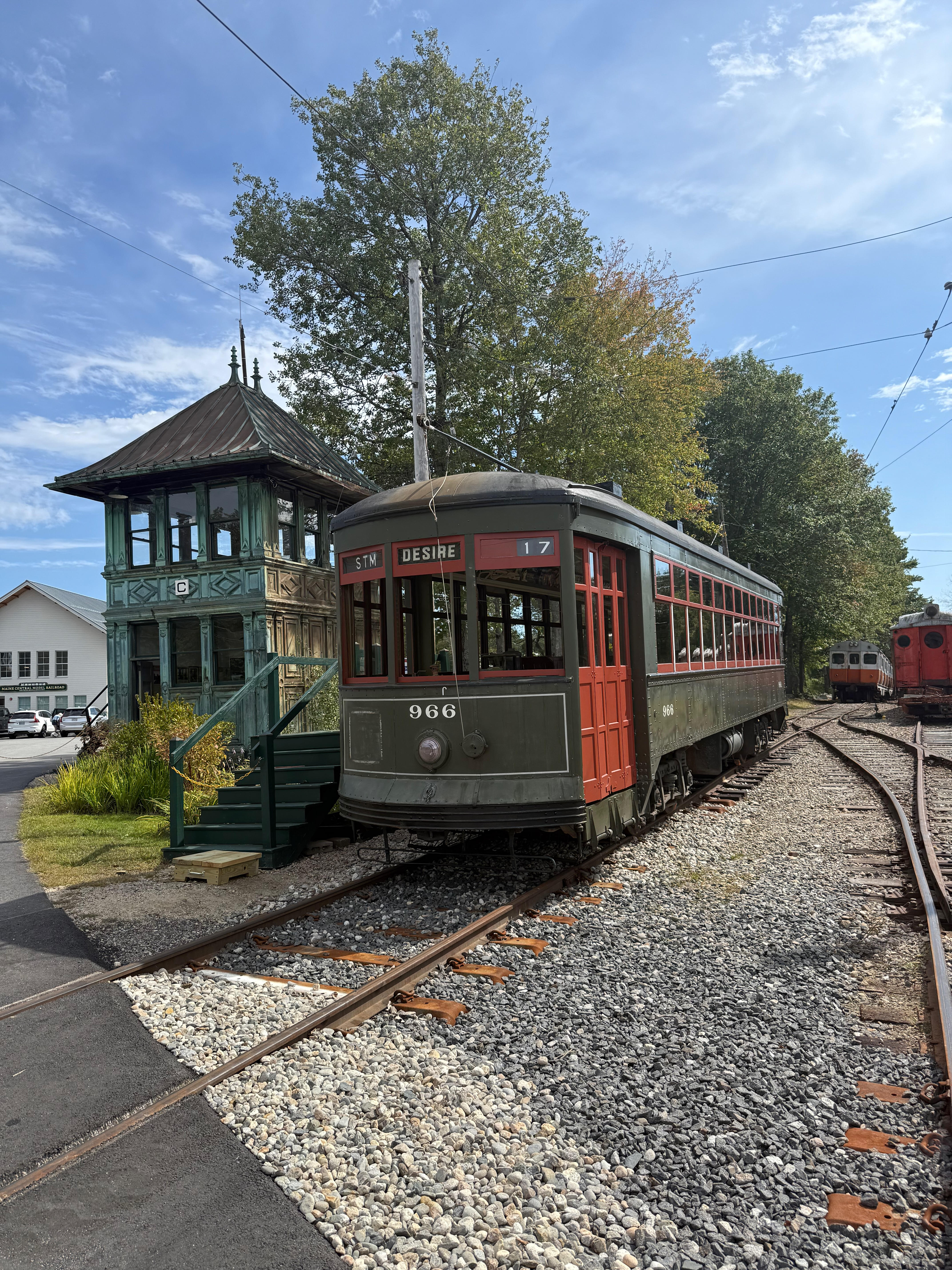 A vintage dark green and red trolley, number 966 with “DESIRE” on its destination sign, is parked on a curved track. To the left stands a historic two-story green wooden signal tower under a blue sky with trees.
