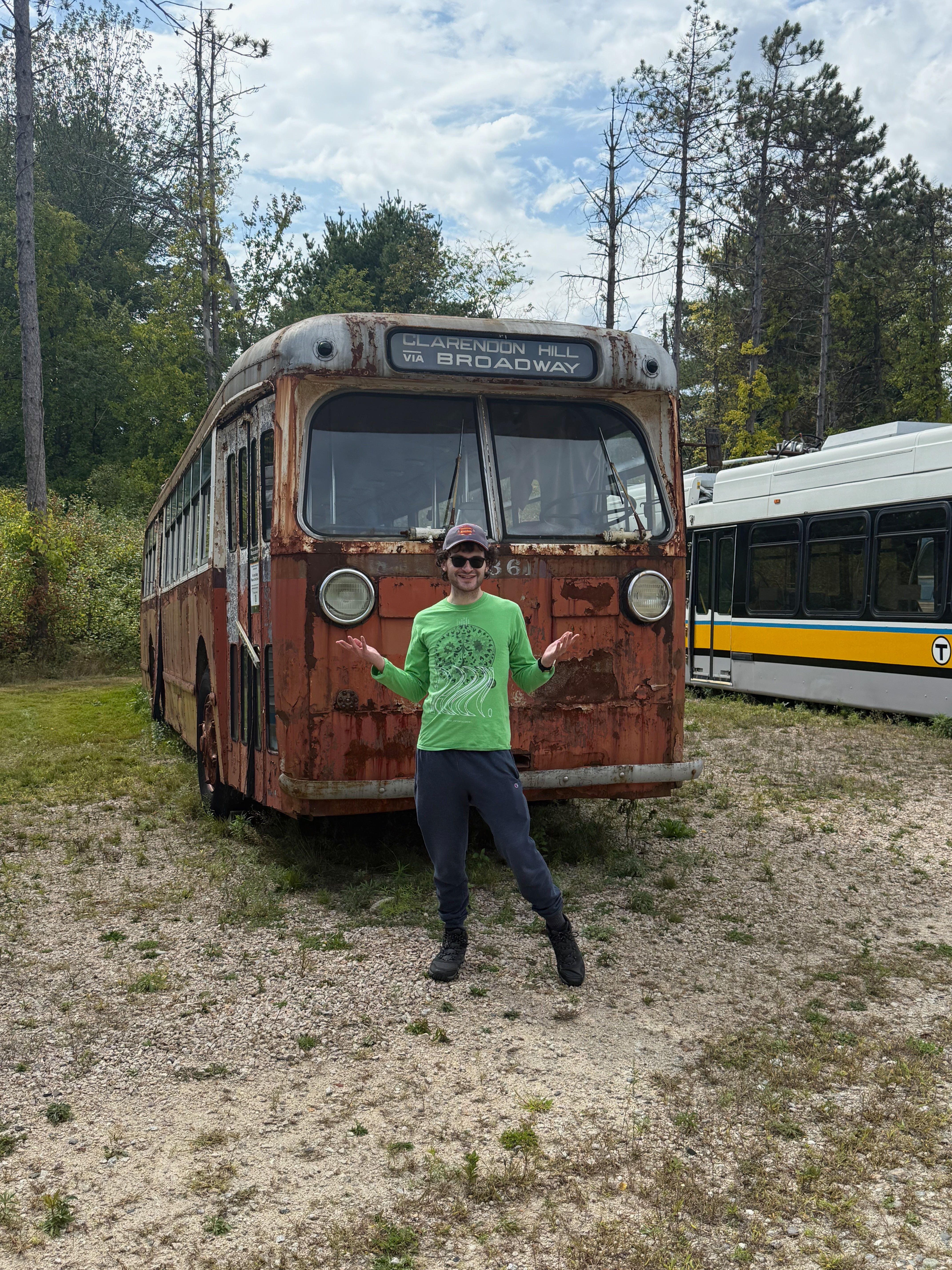Me smiling in front of a very old, rusty red-brown MTA bus. The bus’s destination sign reads “CLARENDON HILL VIA BROADWAY”.