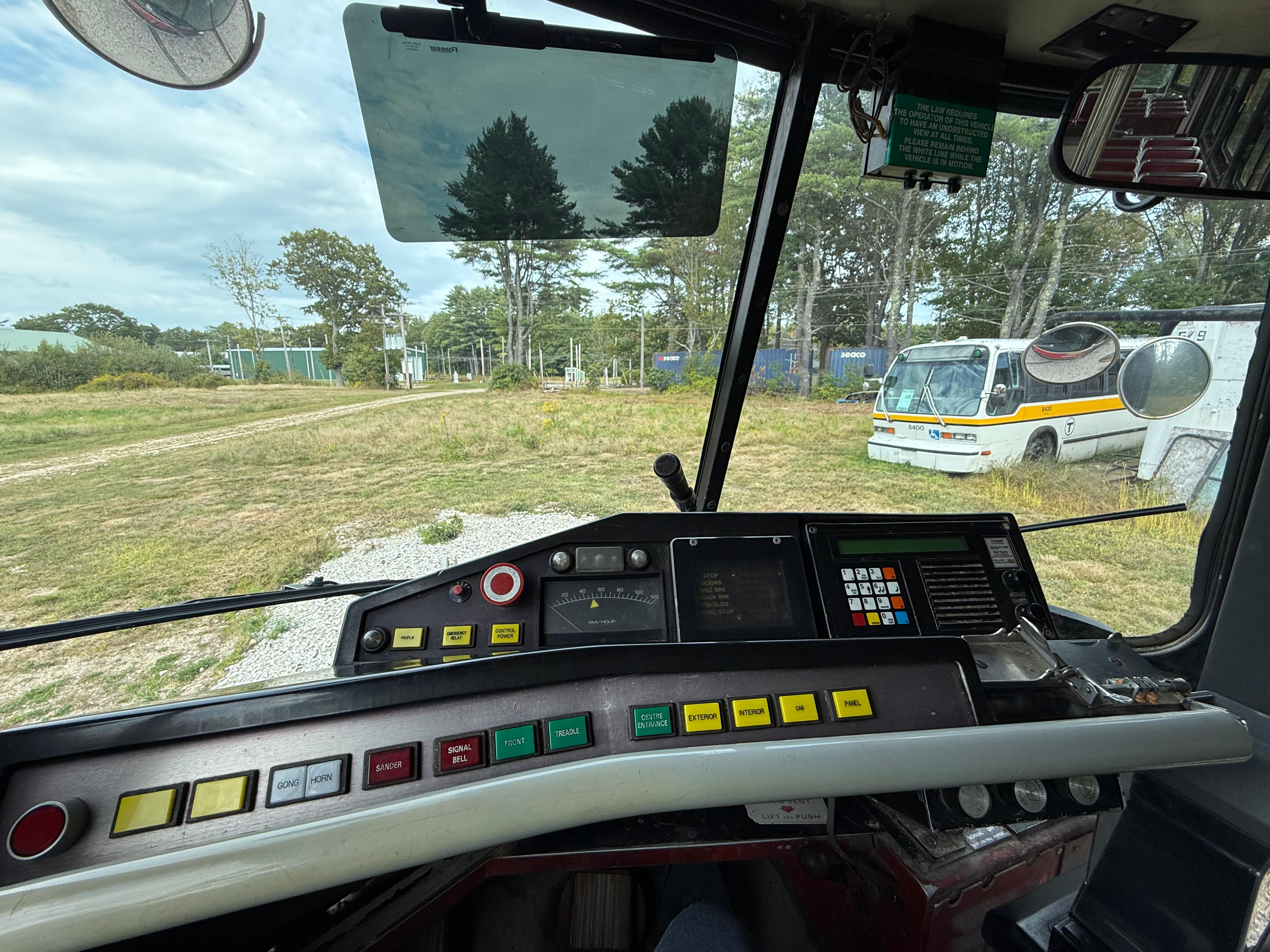 A view from the driver’s seat inside an old TTC Streetcar from Toronto. The dashboard is filled with colorful buttons and analog gauges. Through the large windshield, a grassy field and another bus can be seen in the distance.