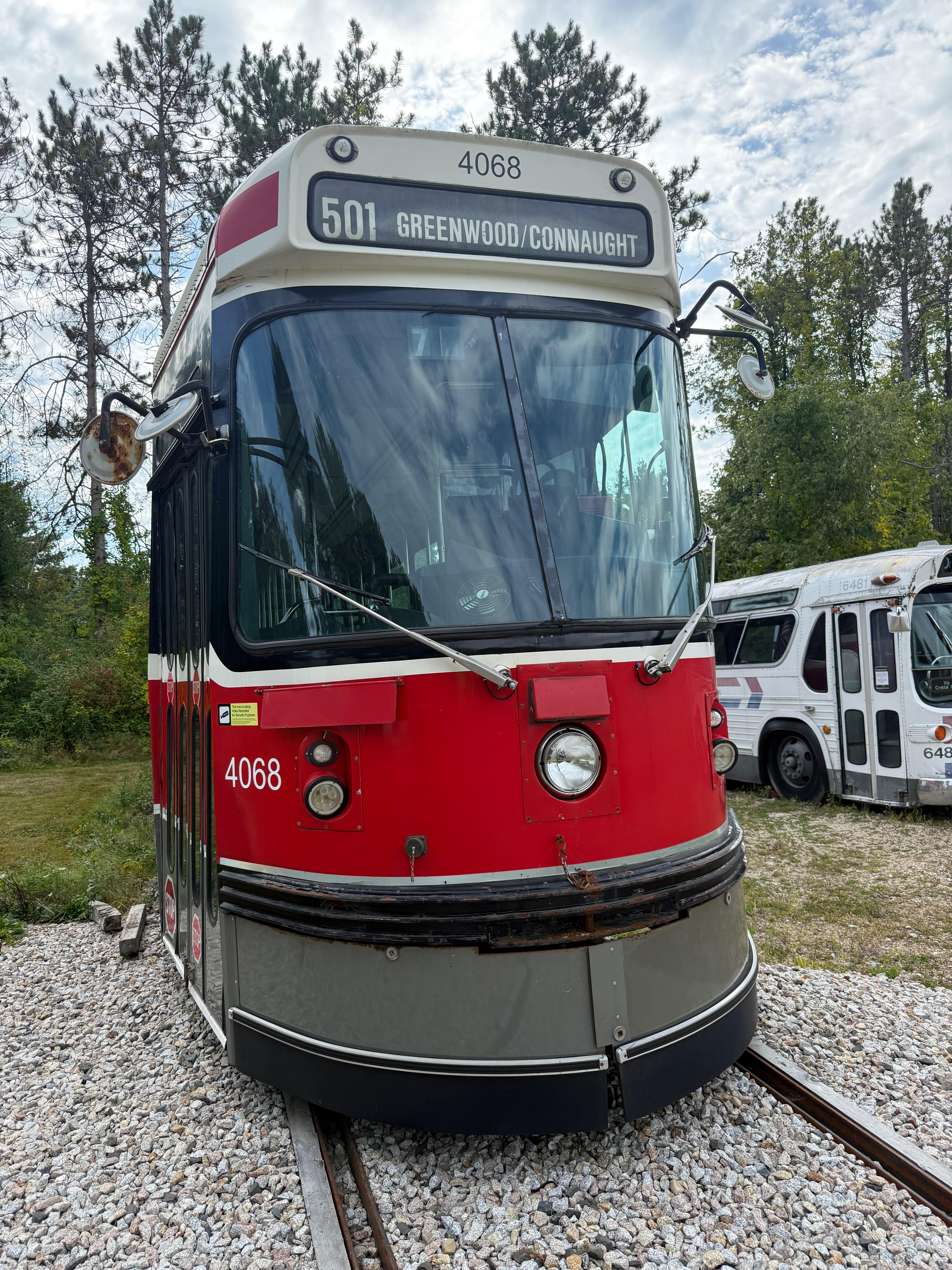 A front-on view of a red, white, and grey Toronto streetcar, number 4068. Its destination sign indicates route “501 GREENWOOD/CONNAUGHT”. The streetcar sits on a gravel track bed with pine trees in the background.