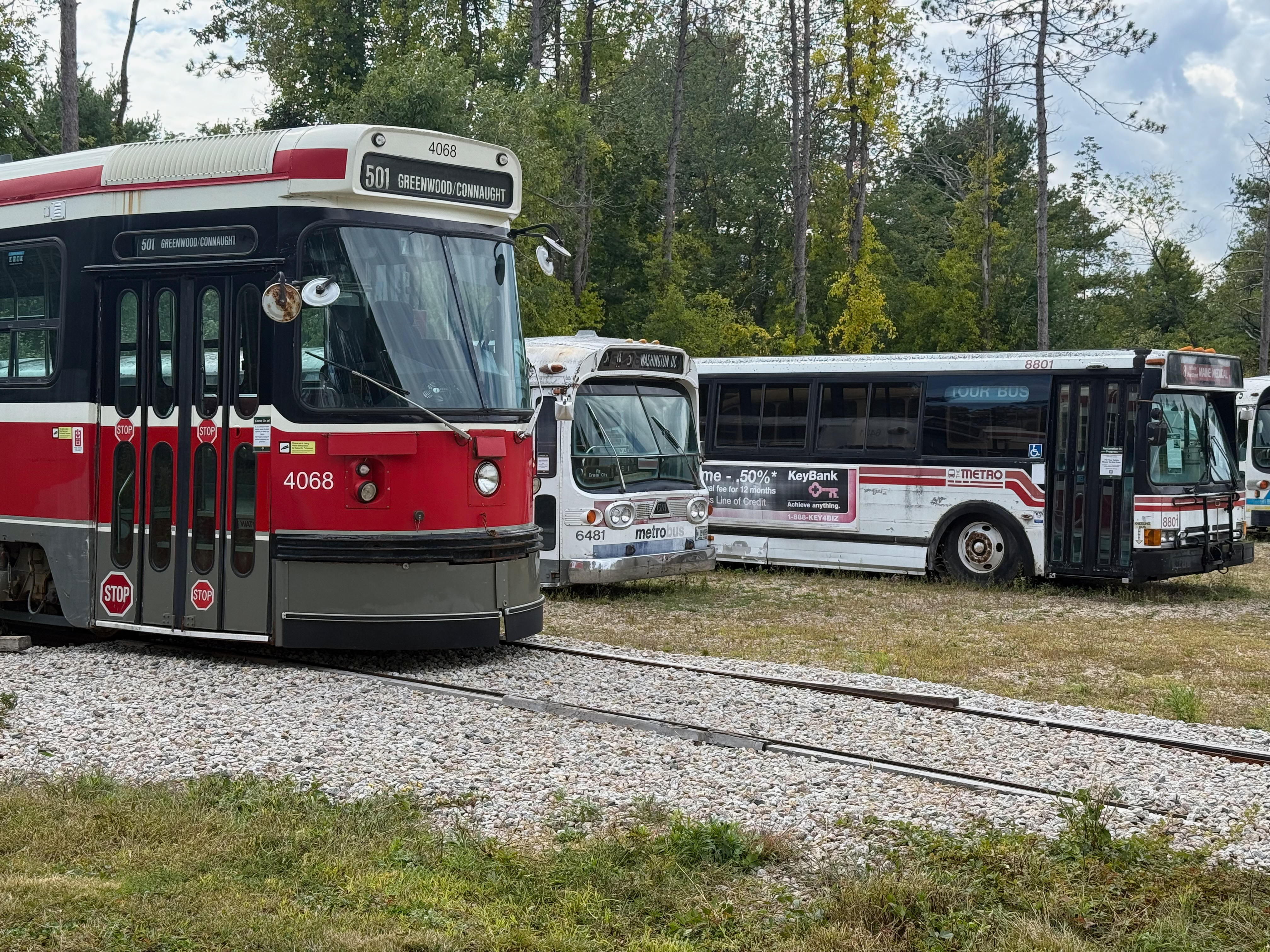 A red and white Toronto streetcar, number 4068, is parked on a track in the foreground. Behind it, on a grassy and gravel area, are two retired white transit buses, including a Washington D.C. metrobus.