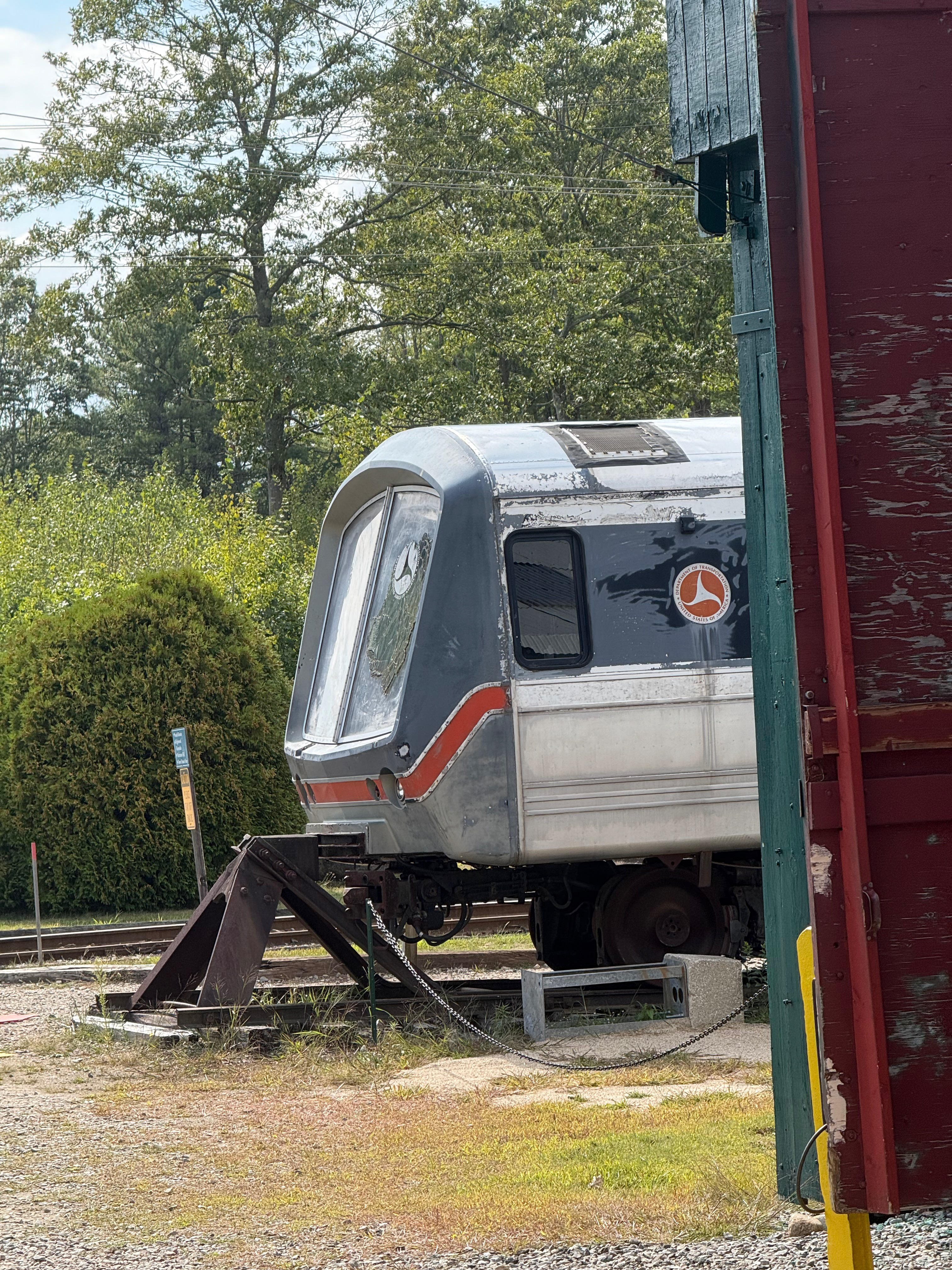 The front end of a sleek, dark grey and silver test subway car parked on a track outdoors. The view is partially framed by a red wooden structure in the foreground.