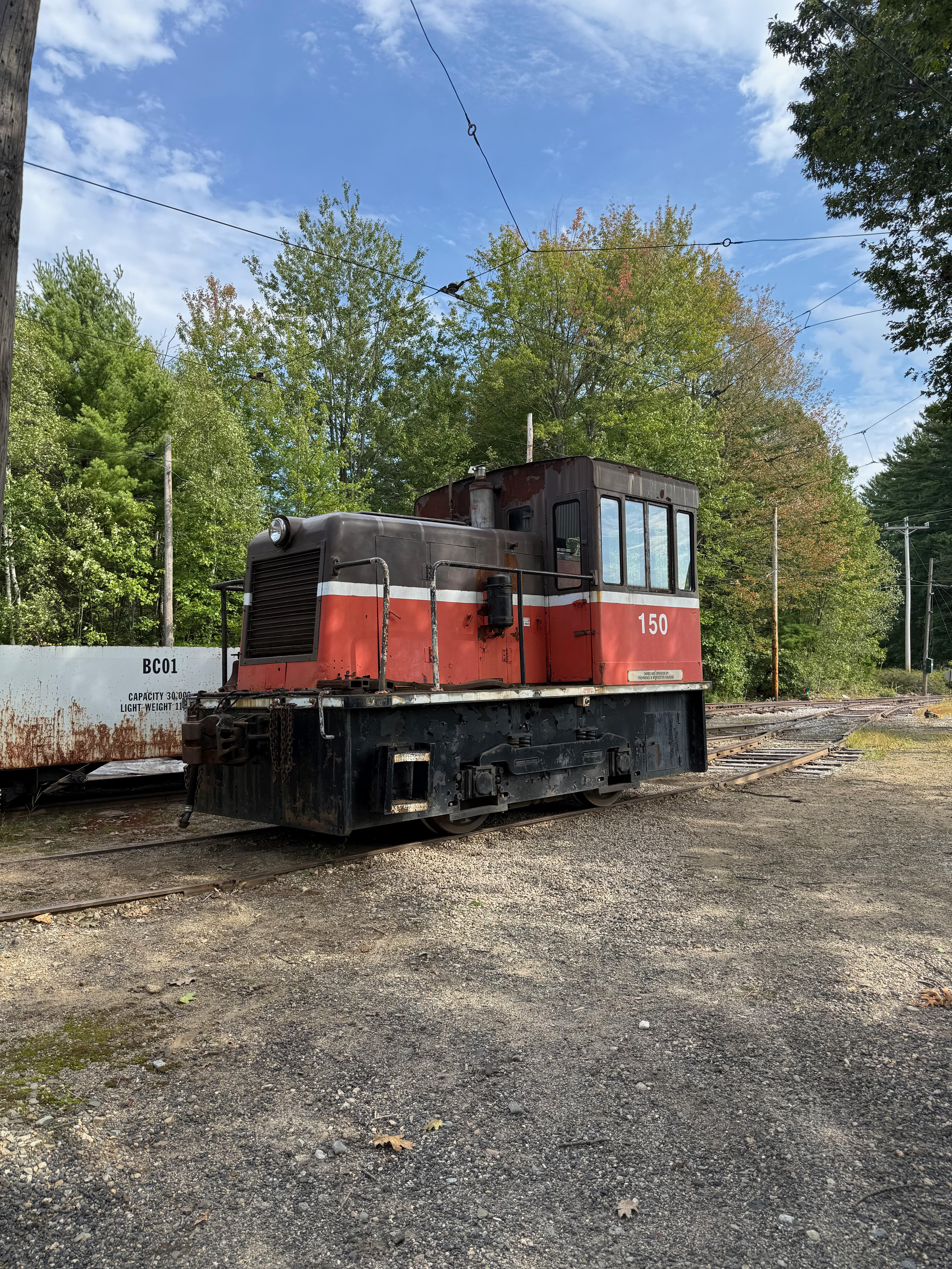 A small red, black, and white industrial switcher locomotive, number 150, sits on a track in a rail yard, surrounded by green trees under a partly cloudy sky.