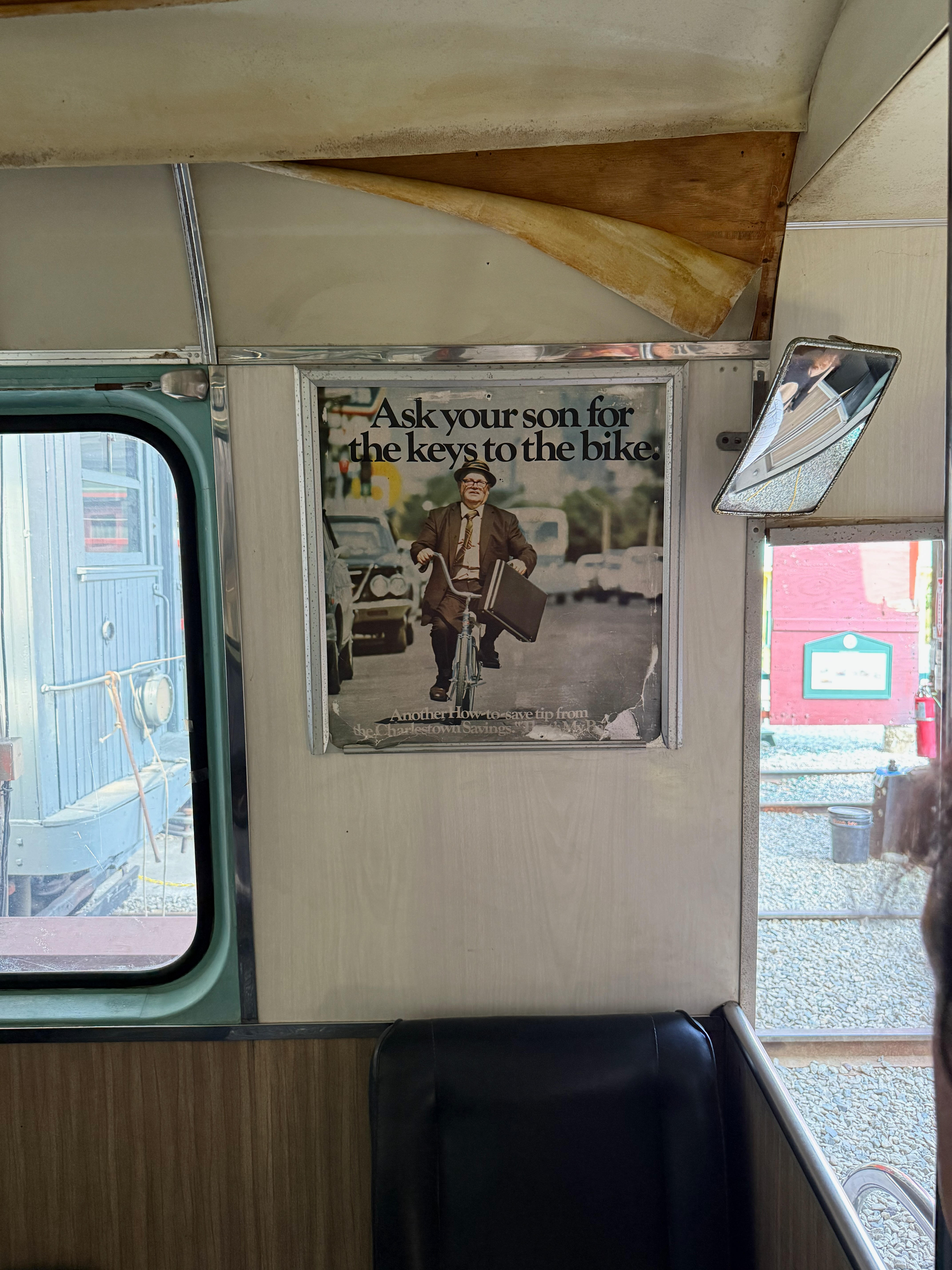 The interior of an old MBTA Trolley Mockup, showing a vintage framed advertisement on the wall. The ad features a man in a suit riding a bicycle with the caption, “Ask your son for the keys to the bike.”