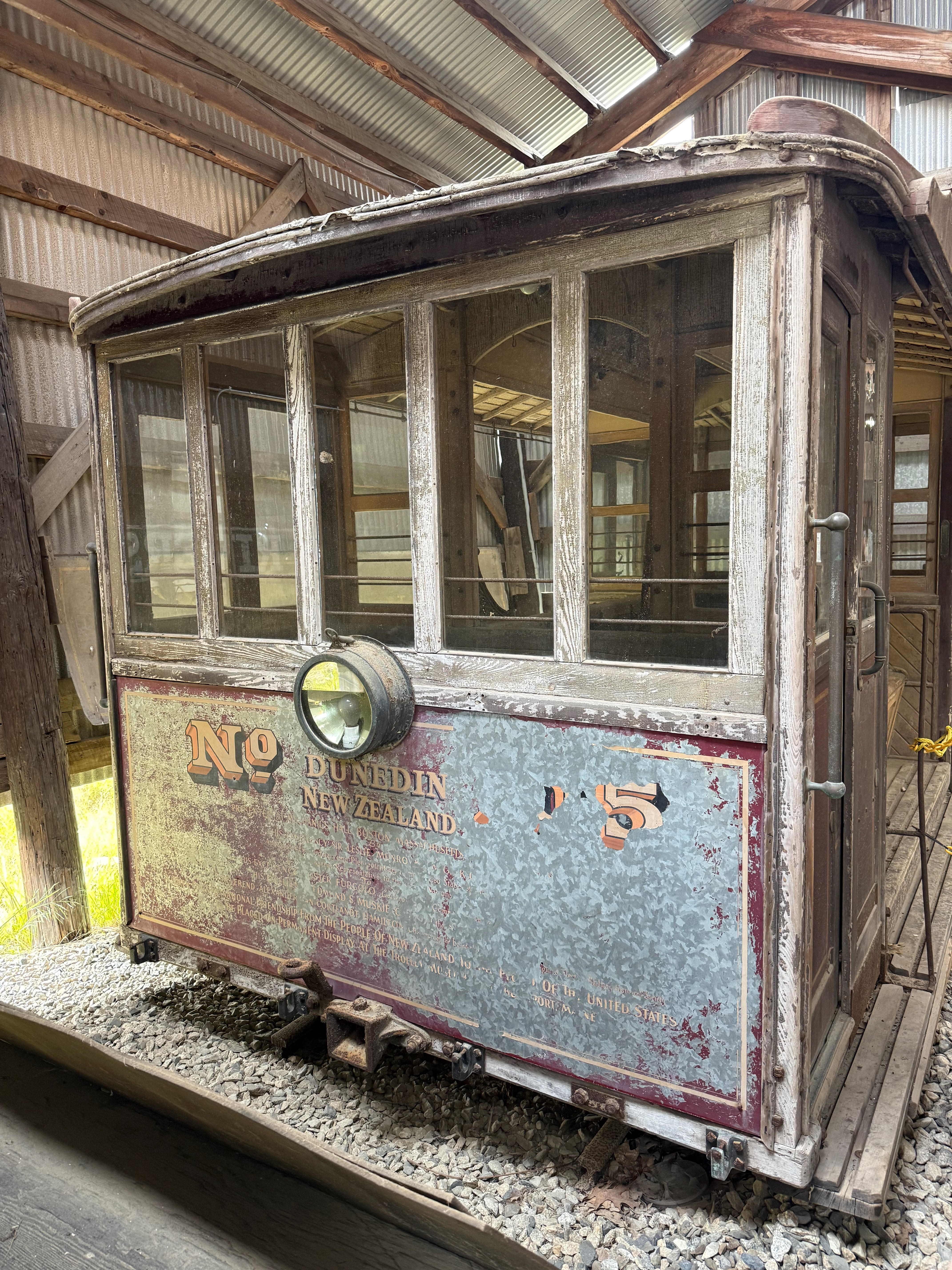 A close-up of a very old, weathered wooden cable car with peeling paint. Faded text on its metal side panel reads “No. 5 DUNEDIN NEW ZEALAND”.