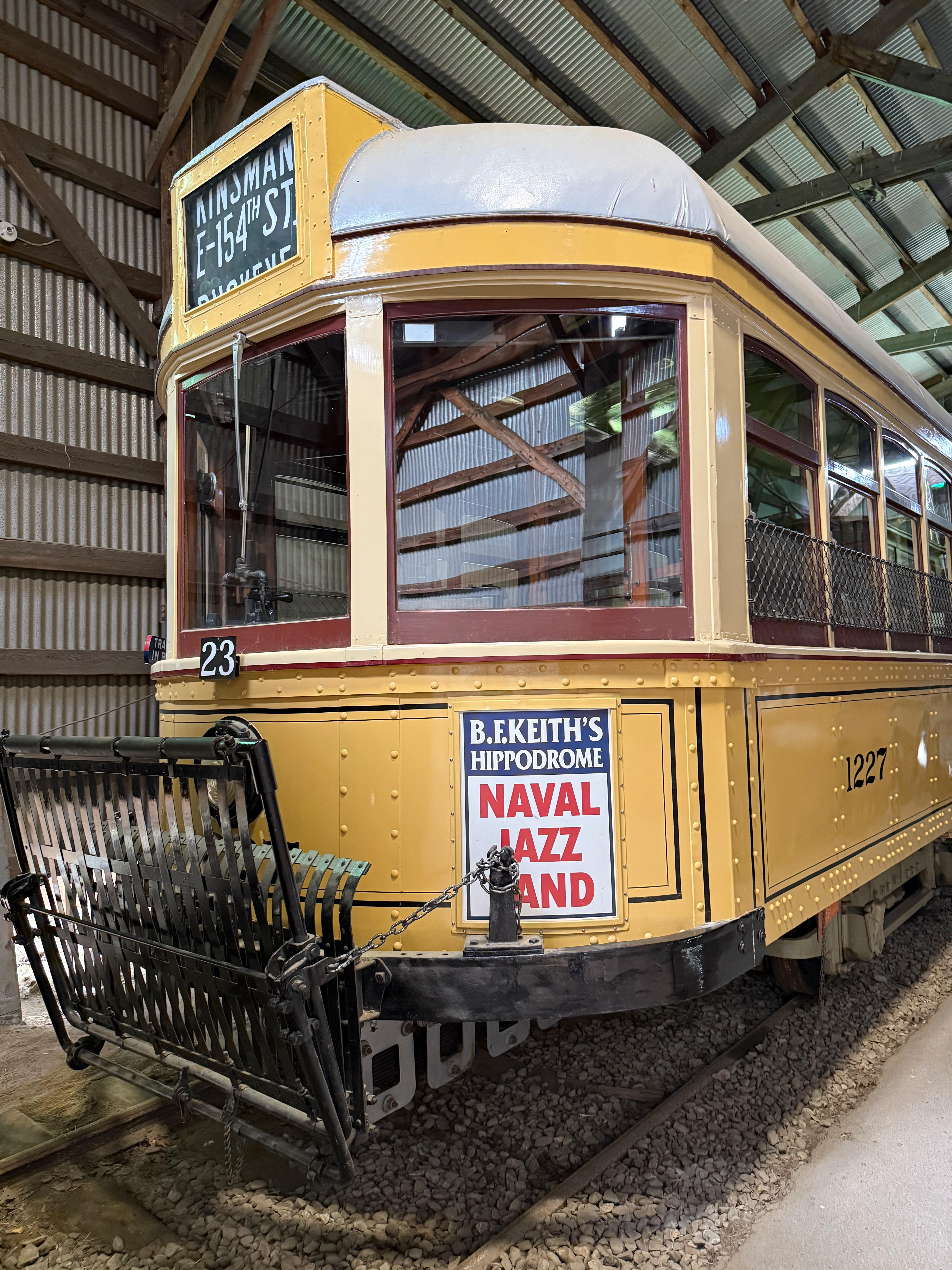 The curved front of a vintage yellow trolley, number 1227, is pictured inside a barn. A sign on its side advertises the “B.F. KEITH’S HIPPODROME NAVAL JAZZ BAND”.