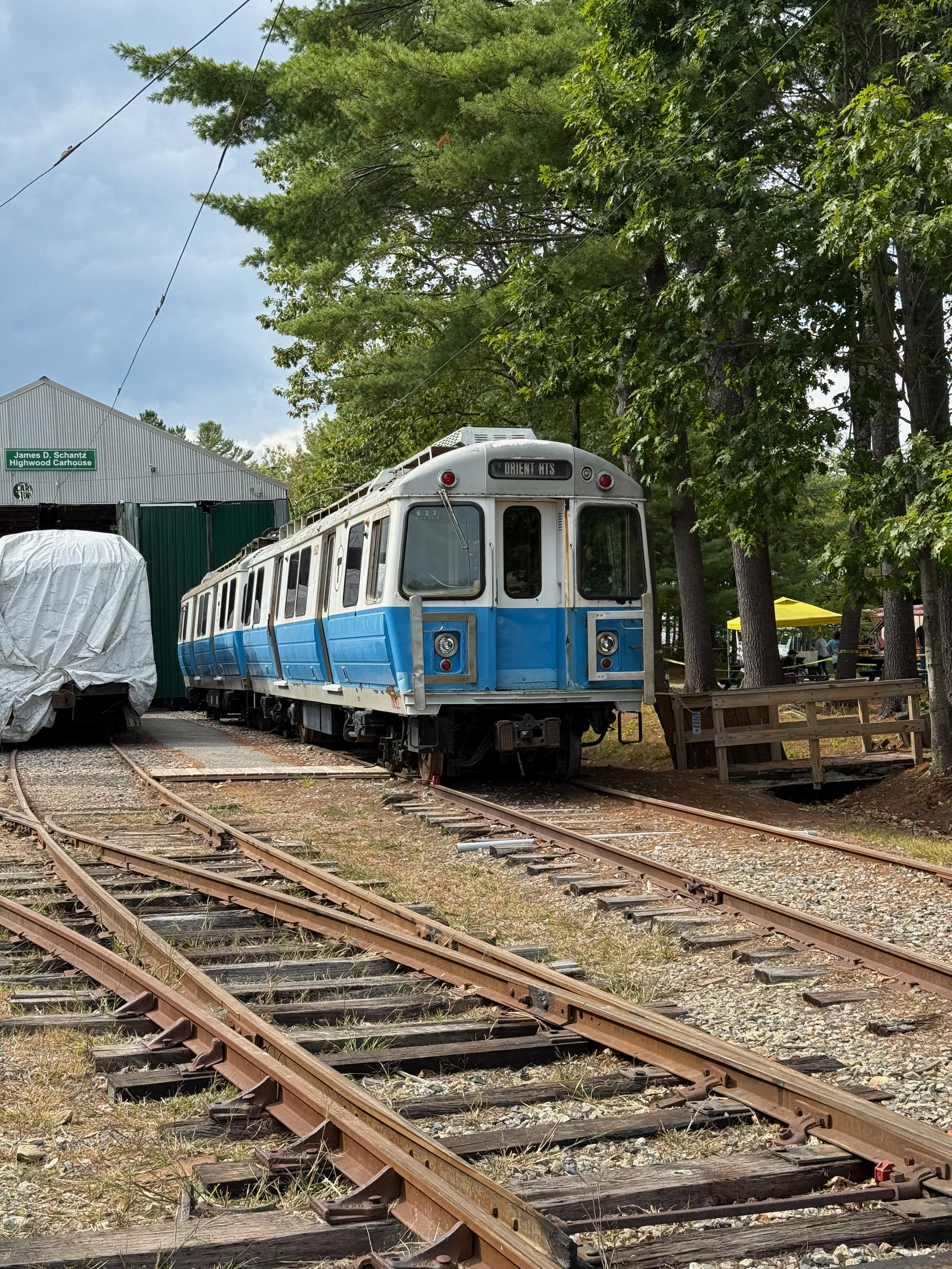 A weathered MBTA Blue Line subway car sits on tracks in a rail yard, with its destination sign set to “ORIENT HTS”. To the left is a green carbarn, and the yard is surrounded by tall trees.