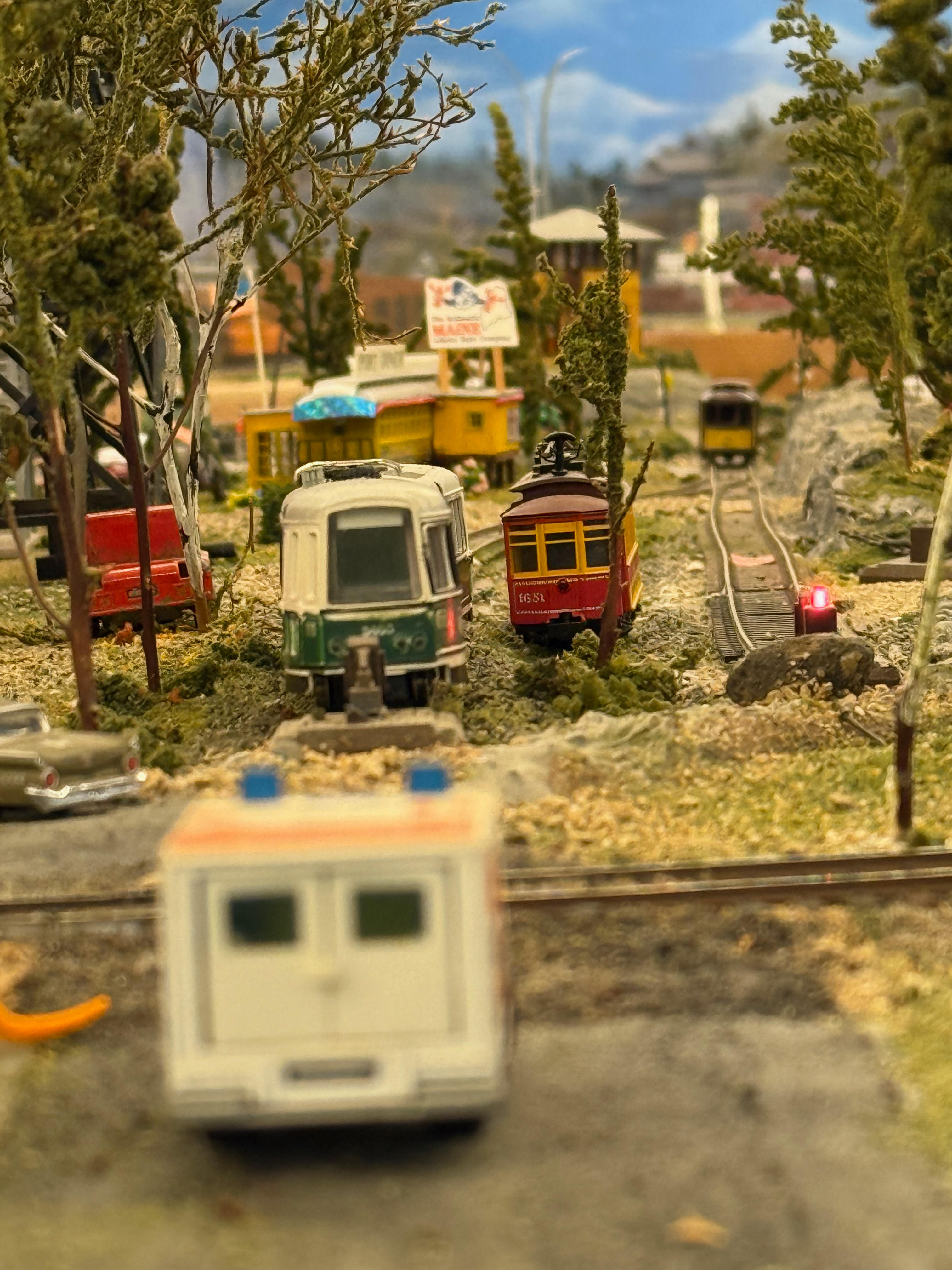 A close-up view of a model trolley scene set in a wooded area. The focus is on a green and white trolley, with other trolleys on different tracks in the background. The back of a blurry model ambulance is in the immediate foreground.