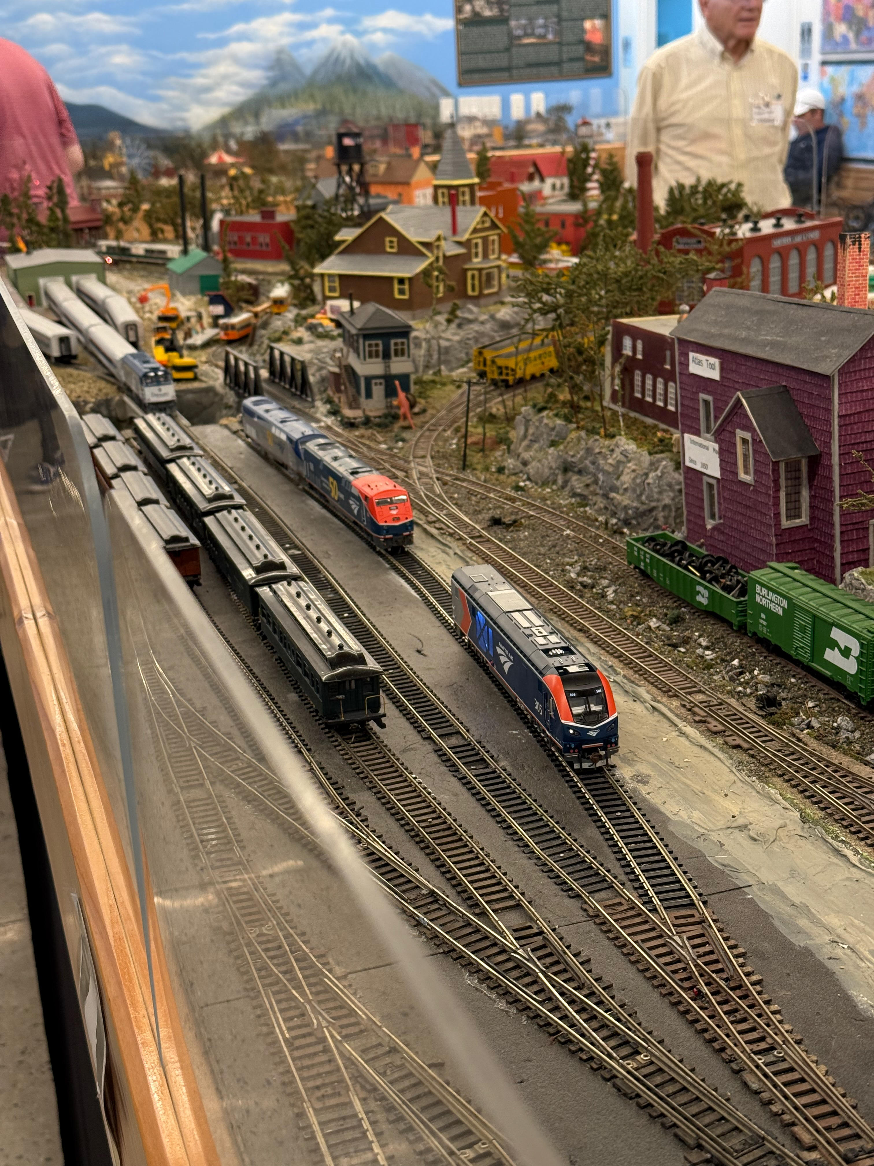 A detailed model train scene with two modern passenger trains on parallel tracks in the foreground: one is a blue and red Amtrak locomotive, and the other is a red and silver MBTA commuter rail locomotive. The background features a miniature town against a painted mountain backdrop.
