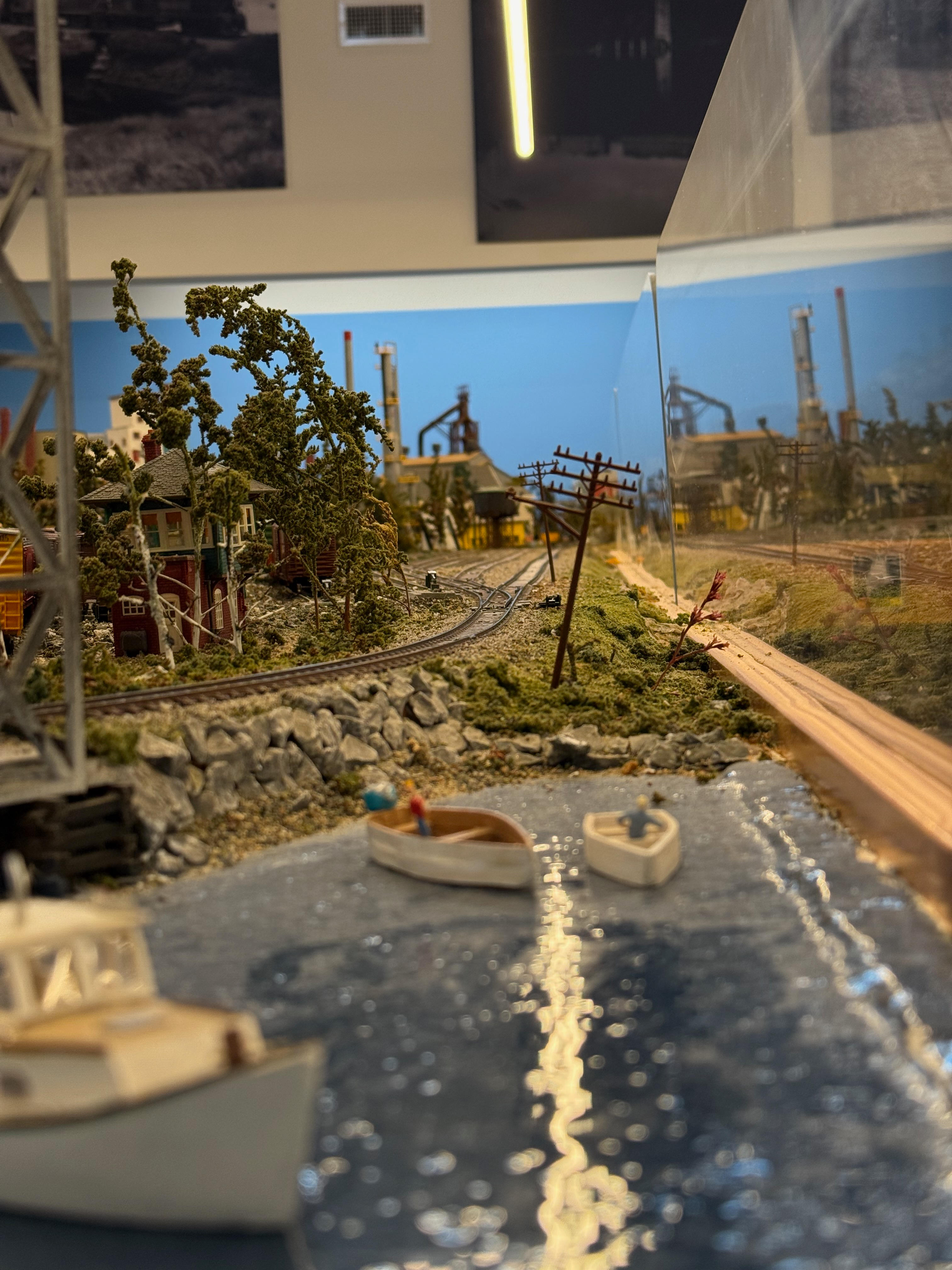 An eye-level shot of a detailed model diorama featuring a body of water with small boats in the foreground. In the background, a train track curves along an embankment towards a model of a large industrial steel mill complex.