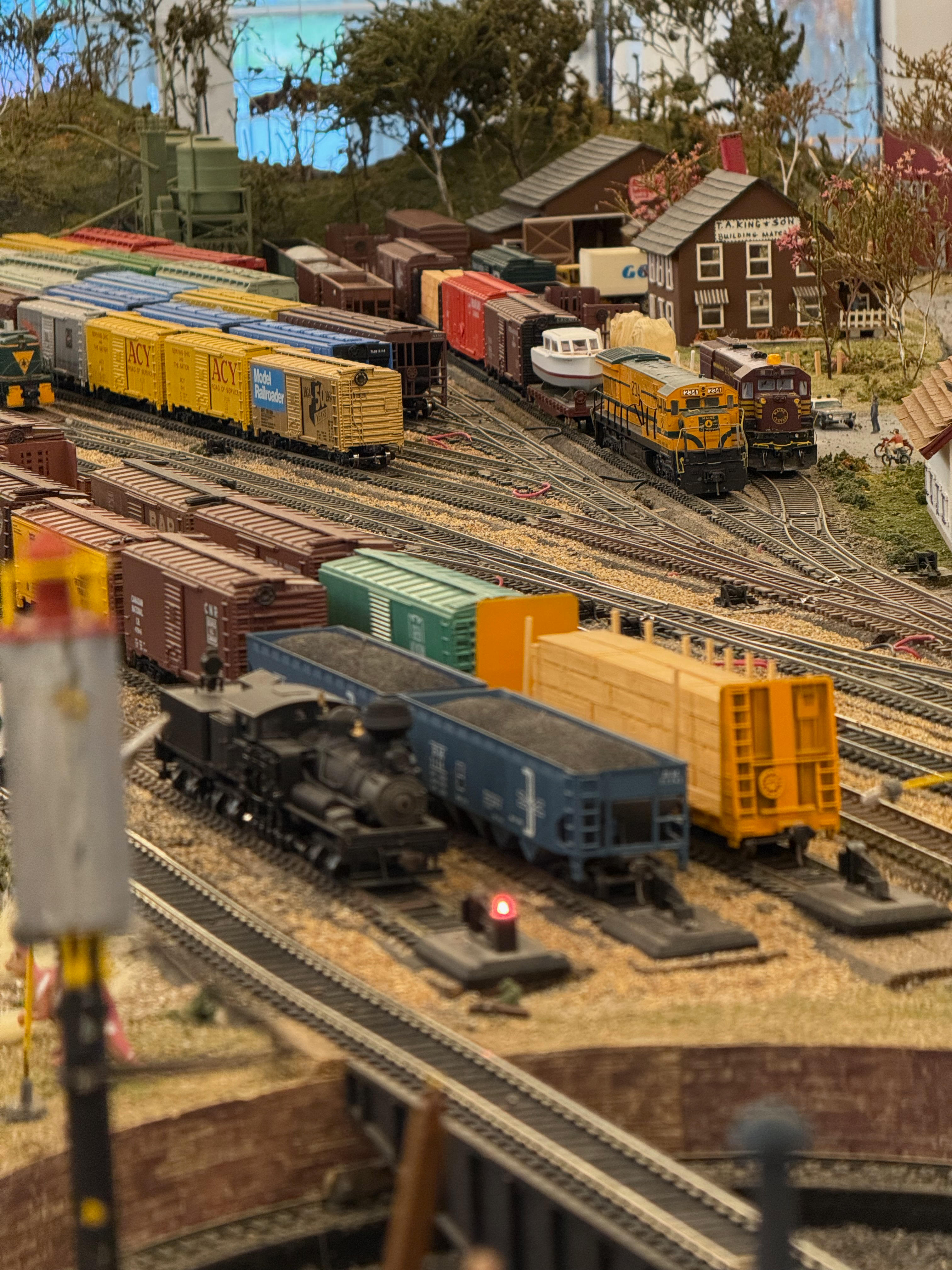A wide, detailed shot of a bustling model railroad yard filled with numerous tracks and a variety of freight trains, including yellow boxcars and a small black steam engine. In the background, diesel locomotives are parked near a small model town.